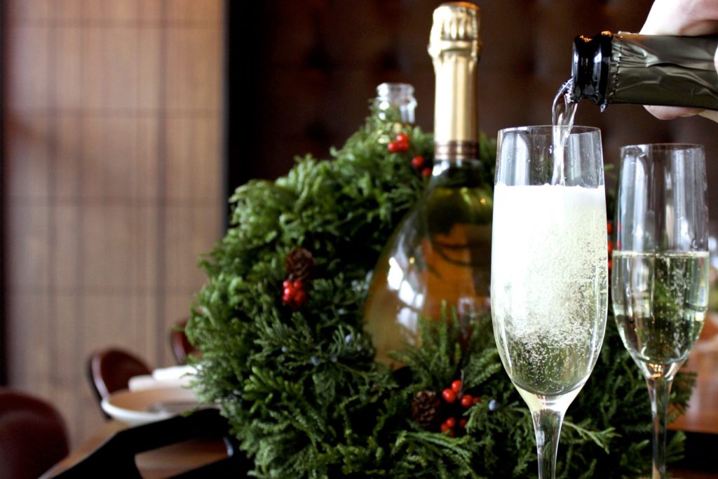 A Christmas wreath around a bottle of wine, and two champagne flutes being filled.