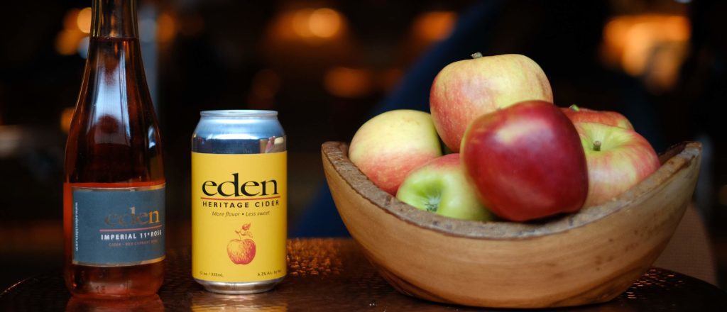 A bottle and can of cider with bowl of apples.