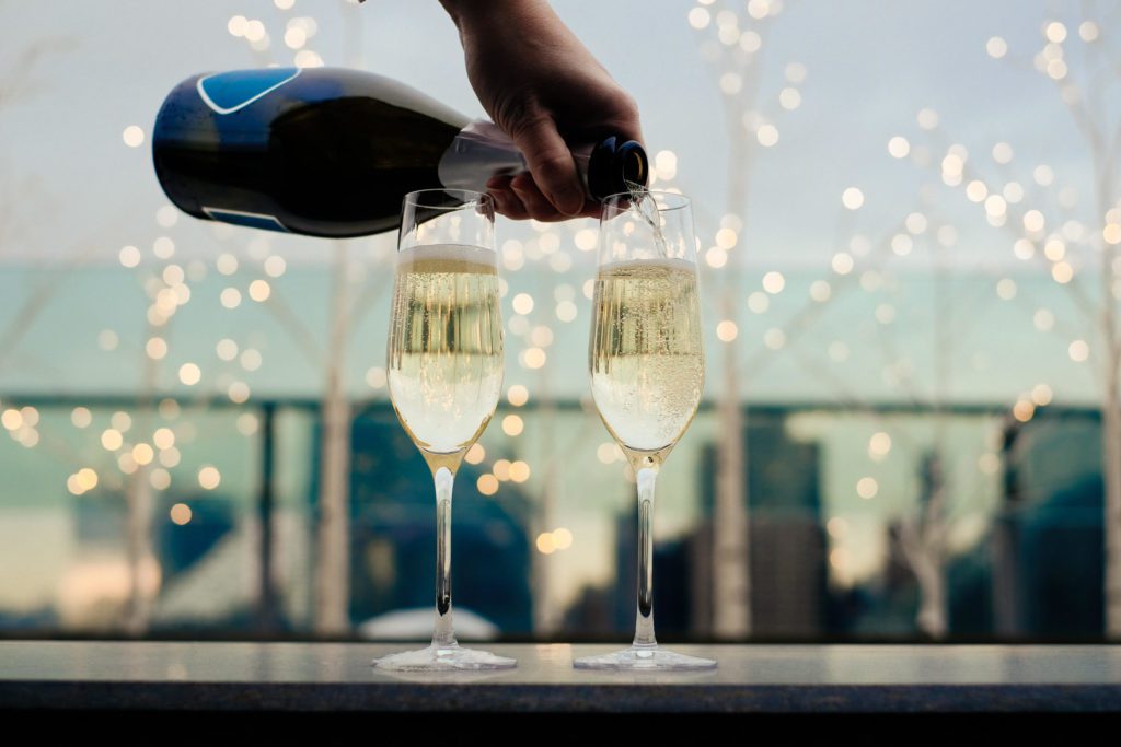 Hand pouring champagne into 2 flutes with twinkle lights and city background.